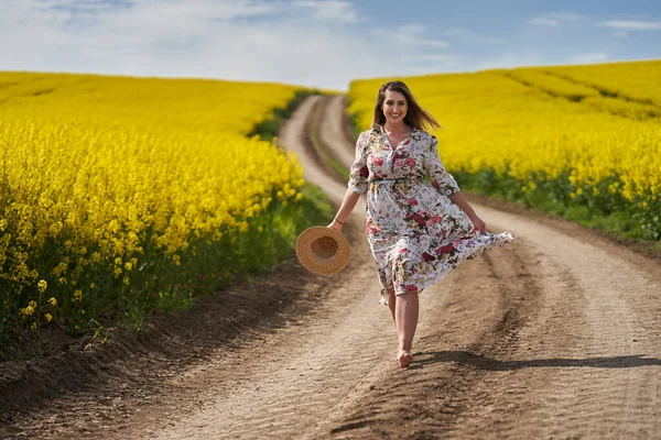 ロマンチックな美しいブロンドのヒスパニック若い女性です花のドレス 裸足で未舗装の道路を通って行く菜の花畑 — ストック写真