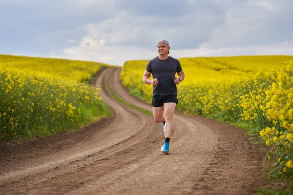 Middle aged distance runner running on a dirt road in a blooming canola field