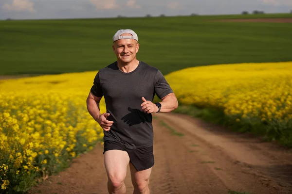在开花结果的油菜地里 中年长跑运动员在泥泞的路上奔跑 — 图库照片