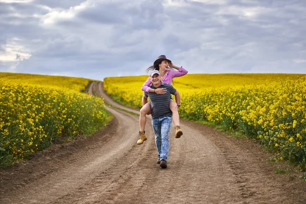 幸せな農家は 彼の妻に彼らの菜の花畑を通って道路上のピギーバックに乗るを与える — ストック写真