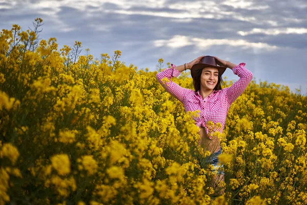 ショートパンツの美しい若いラテン農家の女性 シャツと帽子を果たしました 菜の花畑に立って — ストック写真