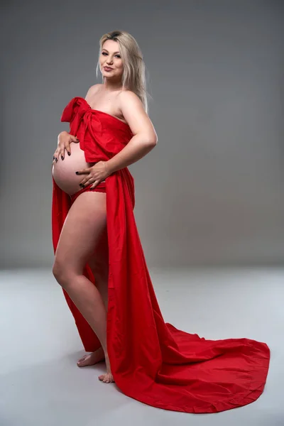年轻美丽的金发孕妇 穿着灰色背景的红色波浪床单 — 图库照片