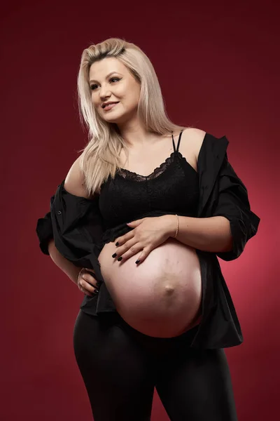 美しいです妊娠中の若いですブロンド女性で黒服 スタジオショットオン赤の背景 — ストック写真