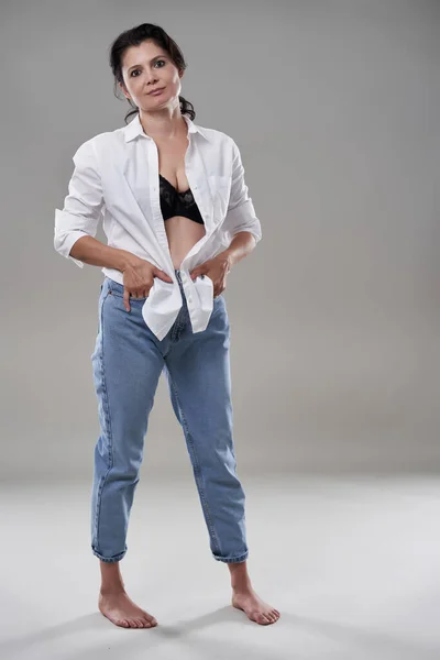 Atractiva Mujer Morena Descalza Camisa Blanca Jeans Sobre Fondo Gris Fotos de stock