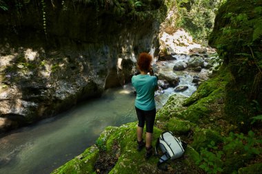 Sırt çantalı bir kadın yürüyüşçü nehir kenarındaki vahşi geçitleri keşfediyor. Kocaman yosun kaplı kayalar.