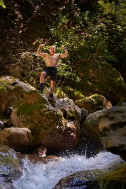 Güçlü üstsüz, atletik bir adam büyük kayalıklarda kaslarını esnetiyor ve bir kanyonda nehir akıntısı var.