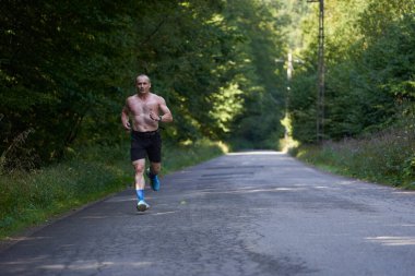 Ormanda asfalt yolda koşan kaslı, atletik bir adam.