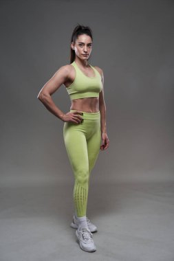 Çekici İspanyol fitness modeli gri arka planda spor kıyafetleriyle poz veriyor. 