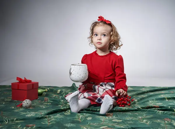 一个可爱的金发卷发小女孩带着一个巨大的圣诞牛奶杯的肖像 在灰色背景下 工作室拍摄 免版税图库图片