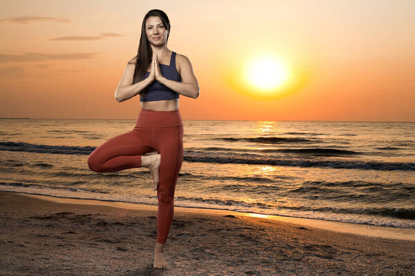 Woman doing yoga on the ocean beach at sunrise with sun in the frame