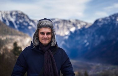 Arka planda karlı dağlar olan kış şapkalı ve ceketli mutlu genç adam, kış portresi