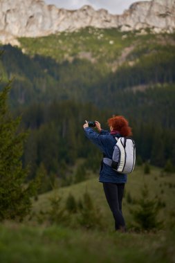 Sırt çantalı bir yürüyüşçü dağlarda cep telefonuyla fotoğraf çekiyor.