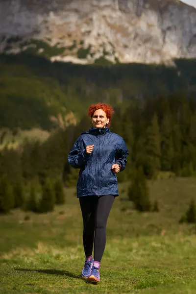 山岳コースの女性トレイルランナー 森林と山の背後にある牧草地で走る ストック写真