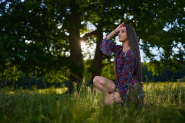 Çiçekli elbiseli güzel bir manken, büyük meşe ağaçlarıyla çayırda poz veriyor. 