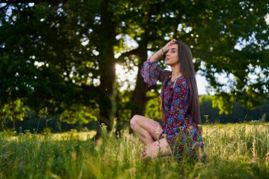 Çiçekli elbiseli güzel bir manken, büyük meşe ağaçlarıyla çayırda poz veriyor. 