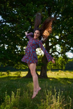 Çiçekli bir elbiseyle, kocaman meşe ağaçları ve ormanları olan bir çayırda sevinçten zıplayıp duran genç ve güzel bir Latin manken.