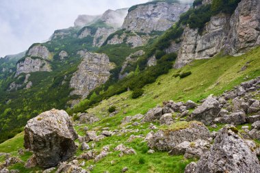 Gün batımından sonra yüksek dağlar sisli bir manzarayla çimenli çayırlar ve büyük kayalarla