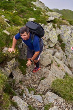 Sıcak bir yaz gününde, kayalık dağlarda zorlu bir patikada sırt çantalı yorgun bir adam yürüyüş yapıyor.