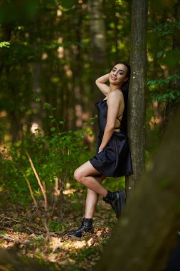 Ormanda duran mini siyah elbiseli çekici Latin kadın.