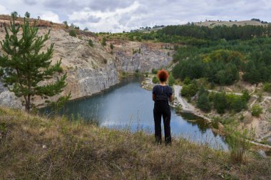Terk edilmiş bir taş ocağında sırt çantasıyla bir gölün etrafında dolaşan kızıl saçlı kadının arka görüntüsü.