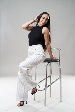 Beyaz pantolonlu ve siyah bluzlu İspanyol kadın gri arka planda sandalyeye yaslanıyor.