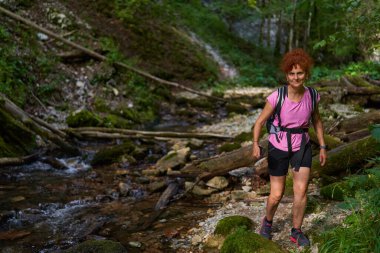 Kızıl saçlı kadın yürüyüşçü gündüz vakti dağlarda nehri geçiyor.