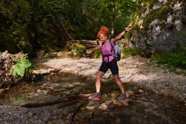 Kızıl saçlı kadın yürüyüşçü gündüz vakti dağlarda nehri geçiyor.