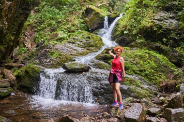 Yazın sonlarında dağlarda şelalesi olan nehir kenarında yürüyüş yapan bir kadın.