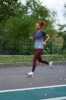Kızıl saçlı kadın sabah parkta koşuyor.