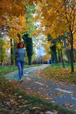 Parkta canlı rengarenk sonbahar ayarı içinde yürüyen kızıl saçlı kadın.