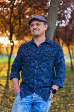 Gün batımında mavi gömlekli ve şapkalı yakışıklı olgun bir adam. Parkta, renkli ağaçlar ve sonbaharda düşen yapraklarla.
