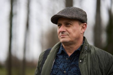 50 'li yaşlarda koyu yeşil ceketli ve şapkalı olgun bir adam parkta.