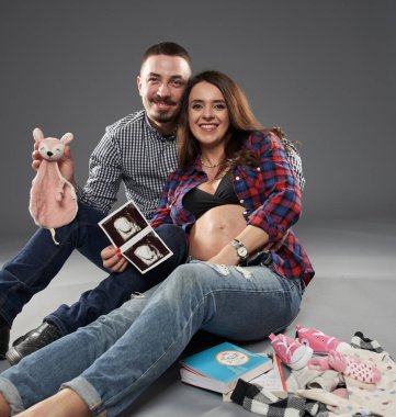 Bebek kıyafetleri, ultrason görüntüleri ve ayakkabılarla yerde oturan, gülümseyen ve bebeklerinin gelişine hazırlanan bir çift.