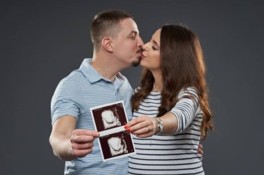 Çiftin, bebeklerinin ultrason görüntülerini tutarken öpüşmelerini beklemek, gri arka plana karşı ebeveynlik yolculuklarını kutlamak,