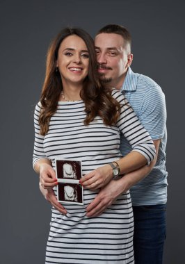 Bekleyen çift, gri arka planda bebeklerinin ultrason görüntülerini tutuyor, heyecanla gülümsüyor ve bekliyor.