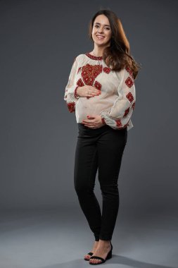 Hamile bir kadın geleneksel işlemeli kırmızı desenli bluz giyiyor, karnını nazikçe tutuyor ve gri arka plana hafifçe gülümsüyor.