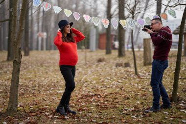 Hamile kadının, seçici bir odak noktasıyla çekilen bayrak altında hamile karısının fotoğraflarını çektiği parkta bir çift bekliyor.