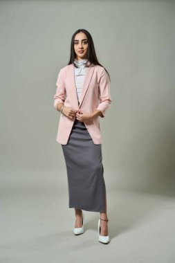 Pembe ceketli, beyaz bluzlu ve gri etekli şık bir kadın yüksek topuklularla rahatça poz verir.