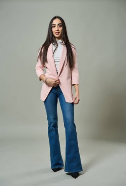 Pembe ceketli, beyaz bluzlu ve parlak pantolonlu güzel kadın yüksek topuklularla rahatça poz veriyor.