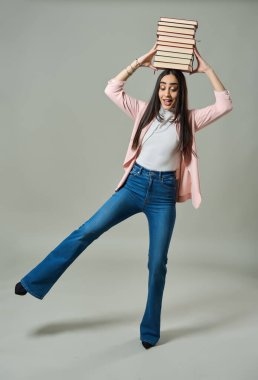 Pembe ceketli şık bir kadın ve kot pantolon bir bacağını kaldırırken kafasında uzun bir kitap yığını dengeliyor.