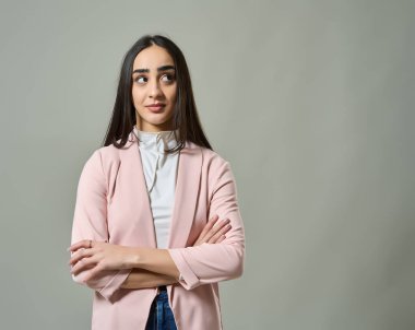 Kolları çapraz duran genç bir kadın ve gri arka plana karşı düşünceli bir ifade. 