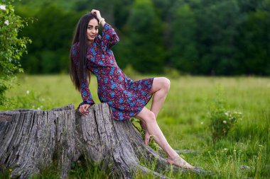 Çiçekli elbiseli genç bir kadın yeşil bir çayırda çıplak ayakla oturmuş, gülümsüyor.