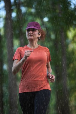 Azimli bir kadın, yağmurda ormanda spor kıyafetleri ve kep takarak koşuyor.