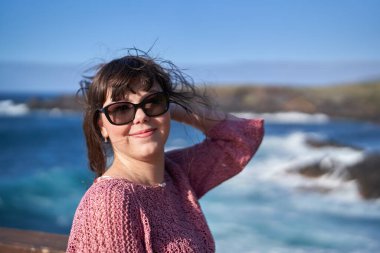 Tenerife 'de okyanusun kenarında gülümseyen güneş gözlüklü ve esintili bir kadın. 
