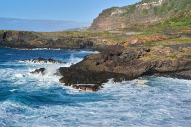 Dalgalar, Tenerife 'de kayalıkları ve yeşil bitkileri olan engebeli bir volkanik kıyıya çarpıyor.