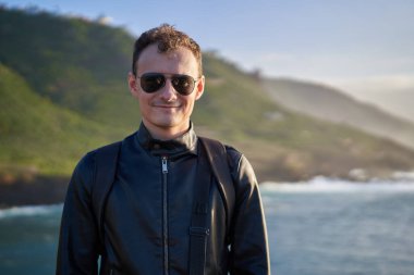 Güneş gözlüklü ve deri ceketli gülümseyen adam gün batımında okyanusun kenarında duruyor ve Tenerif 'in arka planında kayalıklar var.