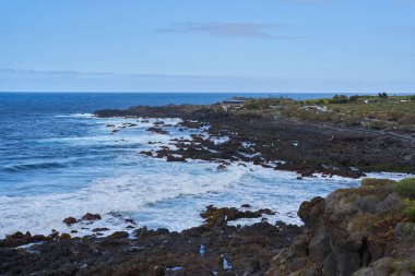 Dalgalar, Tenerife 'de kayalıkları ve yeşil bitkileri olan engebeli bir volkanik kıyıya çarpıyor.