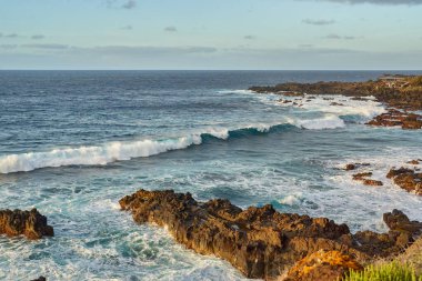Dalgalar gün batımında engebeli kayalarla engebeli bir volkanik kıyıya çarpıyor.