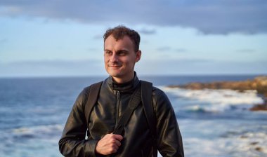 Siyah deri ceketli ve sırt çantalı genç bir gezgin Tenerif 'te kayalık bir kıyıya vuran dalgalarla okyanusun kenarında duruyor.