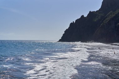 Dalgalar, yükselen kayalıkları ve Tenerife 'deki suyun tadını çıkaran insanların olduğu siyah bir kumsalda süzülüyor.